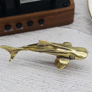 May include: A brass shark-shaped cabinet pull. The shark is detailed with fins, gills, and a tail. The pull is designed to be mounted on a cabinet door or drawer. The Etsy shop name and URL are visible at the bottom of the image.