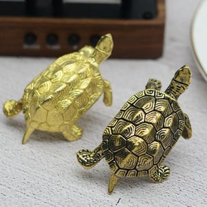 Op de afbeelding: Twee kleine, gedetailleerde messing schildpadfiguren. Eén is helder goudkleurig en de andere is donkerder, antiek goudkleurig. De schildpadden hebben een getextureerd schild en zijn geposeerd met hun poten uitgestrekt. De Etsy shopnaam en URL staan onderaan.