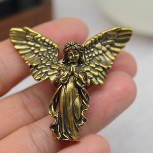Puede incluir: Figura detallada de ángel de latón con alas extendidas y manos juntas. El ángel lleva una túnica fluida y tiene plumas detalladas en las alas. La figura se sostiene en una mano. El texto en la parte inferior dice "Hao DIY" y una dirección web.