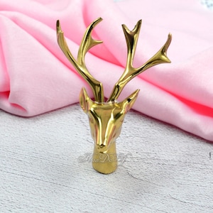 Puede incluir: Figura de cabeza de ciervo dorada con cuernos detallados. La figura tiene un acabado liso y pulido, sobre un fondo de tela rosa. La cabeza de ciervo es un objeto decorativo.