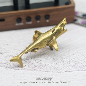 Puede incluir: Figura de tiburón de latón con la boca abierta, que muestra detalles de las aletas y la textura del cuerpo. El tiburón es de color dorado y está sobre un fondo borroso. El texto "HaoDIY" y una dirección web están en la parte inferior.