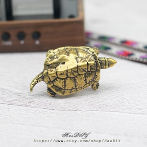 Op de afbeelding: Een kleine, gedetailleerde messing schildpadfiguur. De schildpad heeft een getextureerd schild en zichtbare poten, kop en staart. Het beeldje staat op een wit oppervlak, met een houten object en een patroonstof op de achtergrond. Tekst "HaoDIY" en websiteadres onderaan.