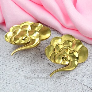 Può includere: Due oggetti decorativi dorati a forma di nuvola. Ogni nuvola ha un design a spirale e un'estremità curva e appuntita. Gli oggetti sono su una superficie chiara con venature del legno e un tessuto rosa sullo sfondo.