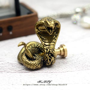 Op de afbeelding: Gedetailleerde messing cobra sculptuur met open mond en uitgespreide kap. De slang is opgerold, met een klein deel van de staart omhoog. De sculptuur is bevestigd aan een cilindrische basis.