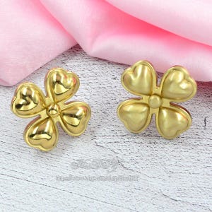 May include: Two gold-coloured, four-leaf clover shaped decorative objects. Each clover has heart-shaped petals and a small, round centre. The objects are on a light-coloured surface with a pink fabric in the background.
