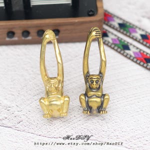 Może przedstawiać: Dwie złote figurki małp. Każda małpa siedzi z uniesionymi ramionami nad głową. Figurki znajdują się na jasnej powierzchni. Nazwa i adres URL sklepu Etsy znajdują się na dole obrazu.