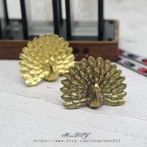 May include: Two gold-coloured peacock-shaped cabinet knobs. The knobs are made of metal and have detailed feather designs. The knobs are on a white wooden surface. The Etsy shop name and URL are at the bottom of the image.