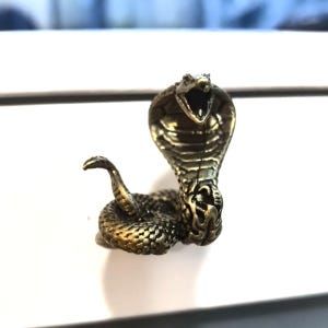 Puede incluir: Figurilla de cobra de bronce detallada con la boca abierta, lista para atacar. Las escamas de la serpiente están intrincadamente detalladas y la figurilla tiene un aspecto vintage y antiguo. La cobra es un símbolo de poder y protección.