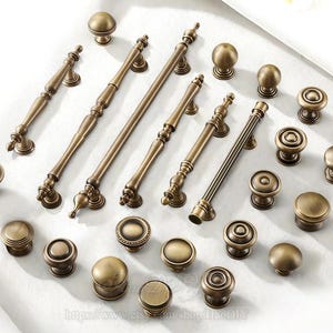 Puede incluir: Un surtido de herrajes de latón antiguo para gabinetes, que incluyen perillas y tiradores. Las perillas son redondas, algunas con detalles decorativos. Los tiradores tienen forma de barra con diseños ornamentados. Todas las piezas tienen un acabado cálido y envejecido.