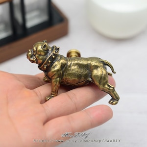 May include: Antique brass bulldog cabinet knob. The dog is depicted in a standing position, wearing a studded collar. The knob has a detailed, textured surface and a warm, aged appearance. A decorative accent for furniture or cabinetry.