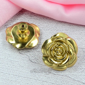 May include: Two brass-coloured rose-shaped cabinet knobs. The knobs have a detailed floral design with a threaded post on the back for easy installation. The knobs are set against a light wood surface and pink fabric.