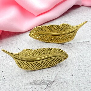 Peut inclure: Deux objets d&eacute;coratifs dor&eacute;s en forme de plumes. Les plumes pr&eacute;sentent des veines d&eacute;taill&eacute;es et une finition brillante. L'arri&egrave;re-plan comprend un tissu rose et une surface blanche.