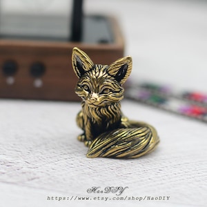 Pode incluir: Estatueta de raposa de latão detalhada com detalhes intrincados de pele. A raposa está sentada com a cauda enrolada em volta do corpo. A estatueta tem uma aparência vintage e antiga. O nome e o URL da loja Etsy são visíveis na parte inferior da imagem.