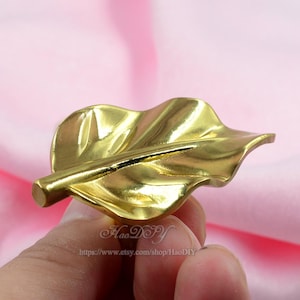 Op de afbeelding: Een goudkleurig, bladvormig decoratief object. Het blad heeft gedetailleerde nerven en een steel. Het object wordt in de hand van een persoon gehouden. De Etsy shopnaam en URL zijn zichtbaar onderaan de afbeelding.