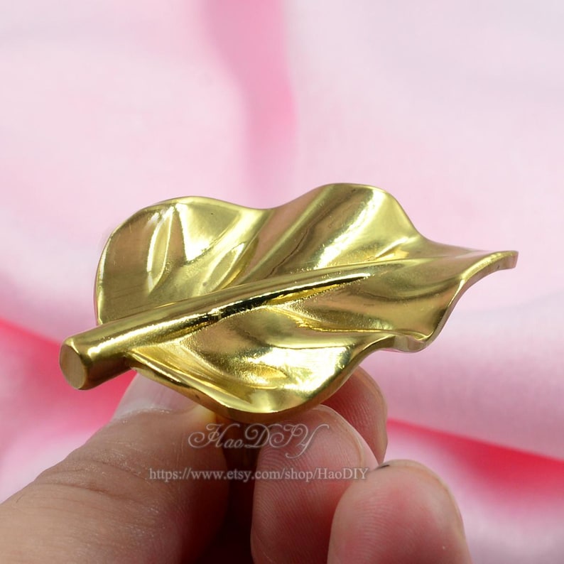 Op de afbeelding: Een goudkleurig, bladvormig decoratief object. Het blad heeft gedetailleerde nerven en een steel. Het object wordt in de hand van een persoon gehouden. De Etsy shopnaam en URL zijn zichtbaar onderaan de afbeelding.