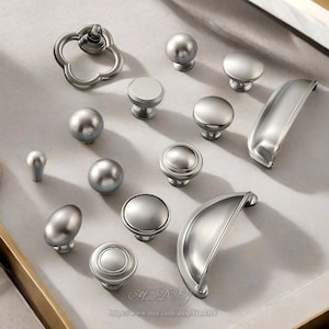 Puede incluir: Una colección de herrajes plateados para armarios, que incluye pomos y tiradores, dispuestos sobre una superficie clara. Los herrajes presentan varios diseños, desde pomos redondos sencillos hasta tiradores más ornamentados. La imagen muestra una variedad de formas y tamaños.