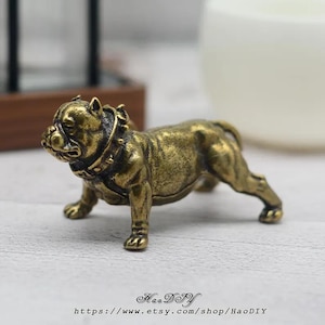 Puede incluir: Pequeña figura de bulldog en bronce, en posición de pie. El perro tiene un collar detallado y una constitución musculosa. La figura está sobre una superficie clara. El nombre y la URL de la tienda Etsy están impresos abajo.