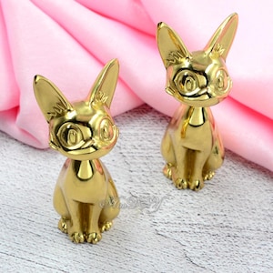 Puede incluir: Dos figuras de gato doradas. Los gatos están sentados con orejas grandes y ojos grandes. Las figuras tienen una superficie brillante y reflectante y están colocadas sobre un fondo rosa.