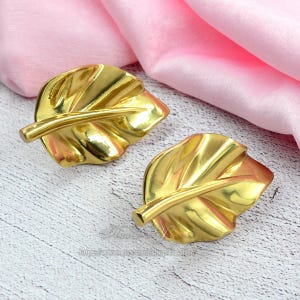 Op de afbeelding: Twee goudkleurige, bladvormige decoratieve objecten. De bladeren hebben gedetailleerde nerven en een steel. De objecten liggen op een lichtgekleurd oppervlak met een roze stof op de achtergrond.