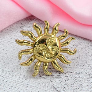 May include: A gold-colored sun-shaped brooch with a smiling face and radiating rays. The brooch has a detailed design and a polished finish, perfect for adding a touch of sunshine to any outfit.