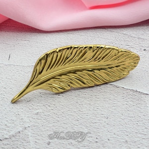 Peut inclure: Un objet d&eacute;coratif en forme de plume de couleur or. La plume pr&eacute;sente des veines d&eacute;taill&eacute;es et une pointe. L'objet est pos&eacute; sur une surface blanche et grise avec un tissu rose en arri&egrave;re-plan. Le texte "Hao DIY" est visible.