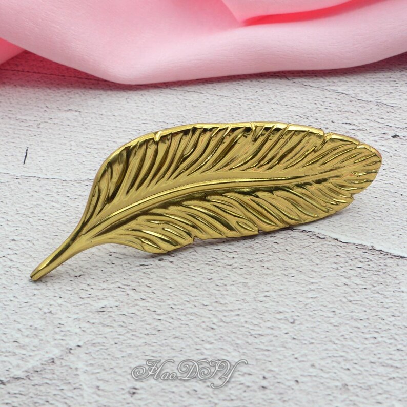Peut inclure: Un objet d&eacute;coratif en forme de plume de couleur or. La plume pr&eacute;sente des veines d&eacute;taill&eacute;es et une pointe. L'objet est pos&eacute; sur une surface blanche et grise avec un tissu rose en arri&egrave;re-plan. Le texte "Hao DIY" est visible.