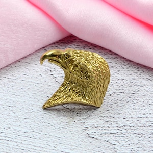 Pode incluir: Uma escultura em metal dourado da cabeça de uma águia. A cabeça da águia é detalhada com penas texturizadas e um bico curvo. A escultura está sobre um tecido rosa e uma superfície de madeira branca.