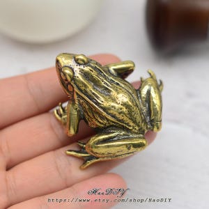 以下が含まれることがあります： テクスチャ加工された肌を持つ、詳細な真鍮製のカエルの置物。カエルはしゃがんだ姿勢で、脚を曲げ、目を前に向けています。置物はヴィンテージ感があります。Etsyショップ名とURLが下部に表示されています。