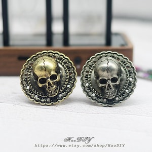 Op de afbeelding: Twee schedelvormige ringen, één goudkleurig en één zilverkleurig, worden getoond. Elke schedel is gecentreerd in een decoratief, cirkelvormig frame. De ringen staan tegen een vage achtergrond. De Etsy shopnaam en URL zijn onderaan zichtbaar.