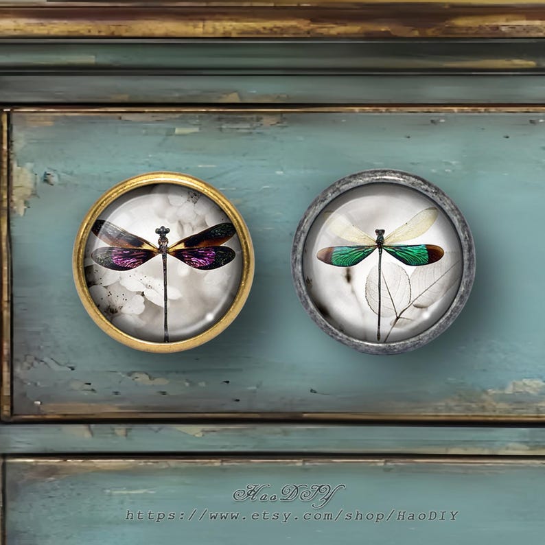 Puede incluir: Dos pomos redondos para armario con dise&ntilde;os de lib&eacute;lulas. Uno tiene un marco dorado y una lib&eacute;lula con alas moradas y negras. El otro tiene un marco plateado y una lib&eacute;lula con alas verdes y blancas.