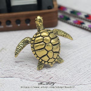 Op de afbeelding: Een gedetailleerd, goudkleurig schildpadbeeldje met uitgestrekte vinnen. De schildpad heeft een getextureerd schild en gedetailleerde kenmerken. De Etsy shopnaam en URL zijn zichtbaar onderaan de afbeelding.