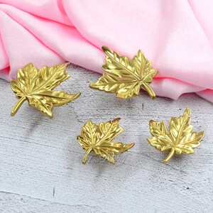 May include: Four gold-colored maple leaf-shaped decorative items. The leaves have detailed veining and are arranged on a pink and white surface. These items could be used as decorative accents or table settings.