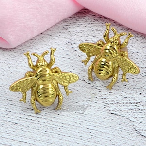 Könnte beinhalten: Zwei goldfarbene, bienenförmige Dekorationsgegenstände. Die Bienen haben detaillierte Flügel, Fühler und Körpersegmente. Es handelt sich wahrscheinlich um dekorative Akzente oder Verzierungen.