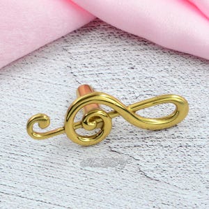 以下が含まれることがあります： 金色のト音記号型のキャビネットノブ。ノブは銅色のステムを持ち、背景にピンク色の布がある明るい色の表面に配置されています。音楽をテーマにした装飾用の装飾要素です。