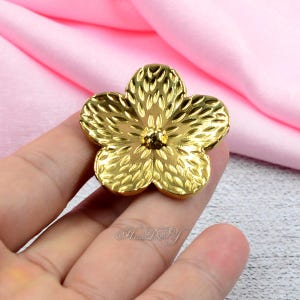 May include: A gold-coloured, five-petal flower-shaped embellishment. The petals have a textured, leaf-like pattern, and the centre features a small, round detail. The flower is held against a pink fabric background.