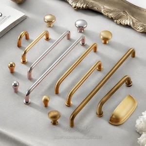 Puede incluir: Una colección de herrajes para armarios en varios acabados metálicos, incluyendo oro, plata y bronce. El surtido presenta pomos, tiradores y un tirador de taza, mostrando diferentes formas y tamaños. El hardware está dispuesto sobre una superficie gris claro.