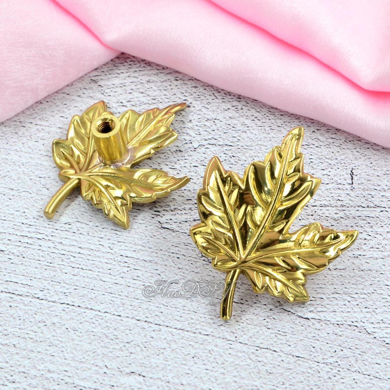 May include: Two gold-colored leaf-shaped cabinet knobs. The knobs have detailed veining and a threaded post for attachment. The leaves are a bright, shiny gold color. The knobs are likely made of metal and are suitable for furniture or decorative projects.