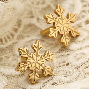 May include: Two gold-colored snowflake cabinet knobs. The knobs are made of metal and have a detailed snowflake design. The knobs are on a white lace background.