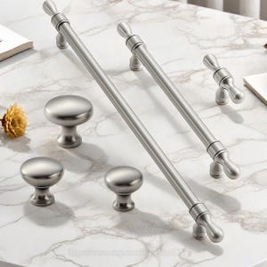 May include: A collection of brushed nickel cabinet hardware, including two long bar pulls, a small pull, and three round knobs. The hardware is displayed on a white and gray marble surface, showcasing a modern aesthetic.