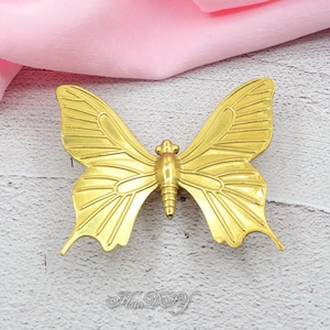 May include: A gold-colored butterfly-shaped decorative object. The butterfly has detailed wing patterns and a textured body. The object is likely made of metal and is displayed on a light-colored surface with a pink fabric in the background.