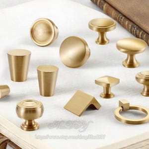 Puede incluir: Una colección de pomos y tiradores de latón para armarios en varias formas y tamaños. El hardware incluye diseños redondos, cónicos y geométricos. Los pomos se muestran sobre una superficie blanca con un libro en el fondo.