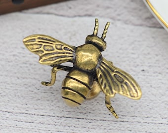 Könnte beinhalten: Ein detaillierter Bienen-Ansteckpin aus Messing im antiken Stil. Die Biene hat strukturierte Flügel und Körpersegmente mit filigranen Fühlern. Der Pin ist ein dekoratives Accessoire, perfekt, um Kleidung oder Accessoires einen Hauch von Natur zu verleihen.