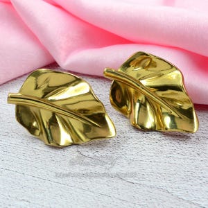 Op de afbeelding: Twee goudkleurige, bladvormige decoratieve objecten. De bladeren hebben gedetailleerde nerven en een glanzend, reflecterend oppervlak. De objecten staan tegen een roze stoffen achtergrond.