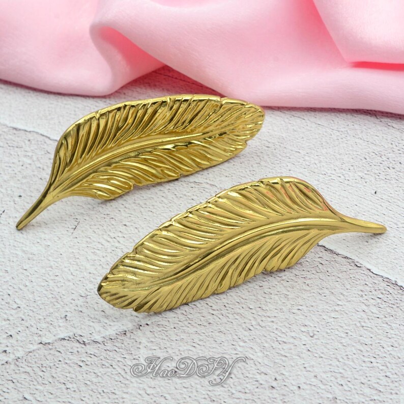 Peut inclure: Deux objets d&eacute;coratifs en forme de plumes de couleur or. Les plumes pr&eacute;sentent des veines d&eacute;taill&eacute;es et une finition lisse et polie. Les objets sont pr&eacute;sent&eacute;s sur une surface claire avec un tissu rose en arri&egrave;re-plan.