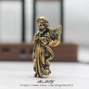May include: A detailed, antique-style brass angel figurine with folded hands and feathered wings. The angel is wearing a flowing gown and has detailed facial features. The Etsy shop name and URL are visible at the bottom of the image.