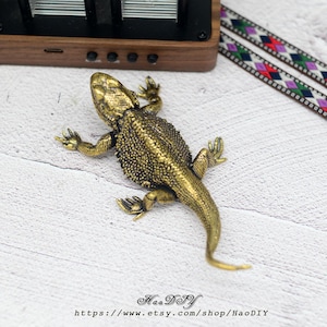 Puede incluir: Figurita de lagarto de latón de estilo antiguo y detallado. El lagarto está posado con las patas extendidas, mostrando una piel texturizada y detalles intrincados. La figurita está sobre una superficie blanca, con un objeto de madera y una cinta estampada en el fondo.