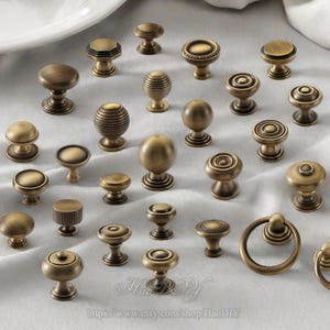 Könnte beinhalten: Eine Sammlung antiker Messing-Schrankknöpfe und -Griffe in verschiedenen Designs. Die Beschläge umfassen runde, kugelförmige und geometrische Formen mit einem warmen, gealterten Finish. Die Knöpfe und Griffe sind auf einer hellen Oberfläche angeordnet.