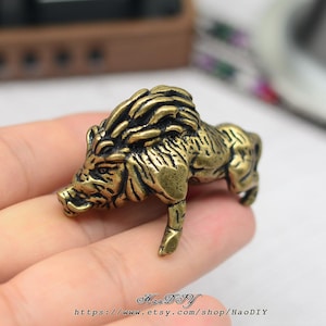 Puede incluir: Una detallada figura de latón de un jabalí, que muestra texturas intrincadas y un acabado envejecido. La escultura representa al animal en una pose de caminar, con colmillos visibles y una melena texturizada. El nombre y la URL de la tienda Etsy están en la parte inferior.