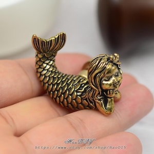 Könnte beinhalten: Eine bronzefarbene Meerjungfrau-Figur mit detaillierten Schuppen und fließendem Haar. Die Meerjungfrau liegt in einer liegenden Pose und stützt ihren Kopf auf ihre Hand. Der Schwanz ist gebogen, und das Gesamtdesign ist aufwendig und dekorativ.