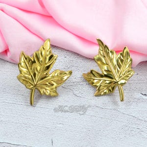 May include: Two gold-colored maple leaf-shaped decorative accents. The leaves have detailed veining and a stem. The background includes a pink fabric and a white and gray surface.
